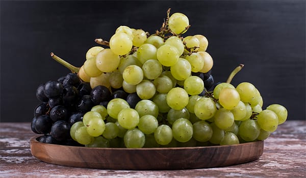 Green and black grapes piled on a wooden platter against a dark background.